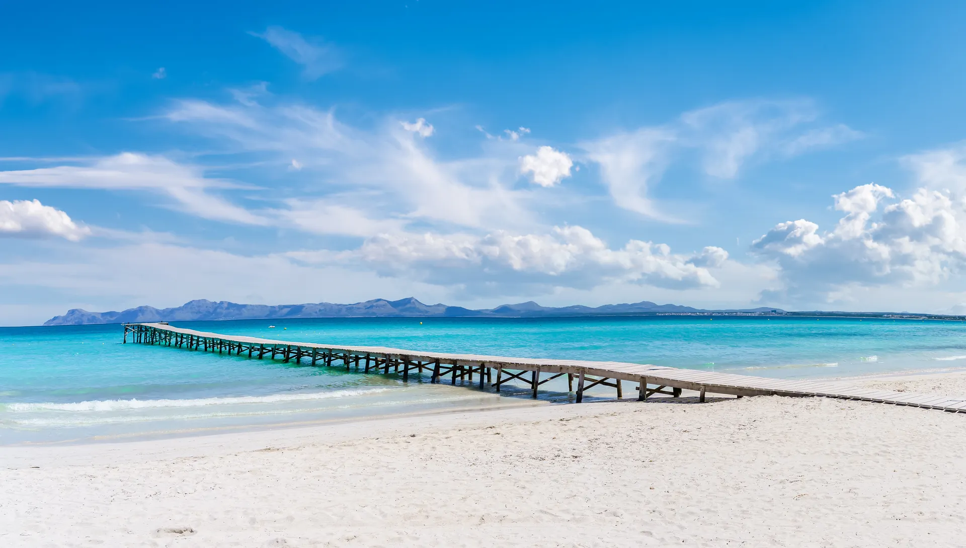 Langer Holzsteg an einem weißen Sandstrand auf Mallorca, türkisfarbenes Meer und Berge am Horizont