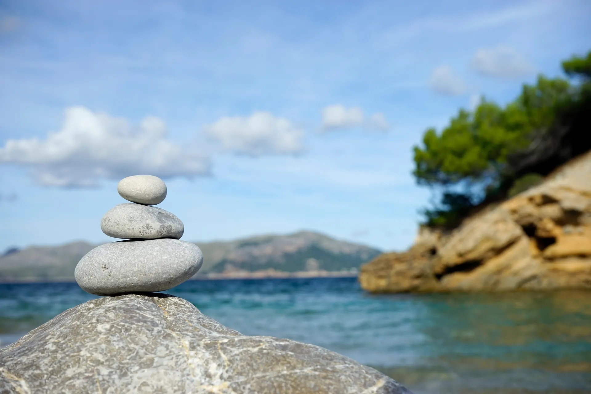 Three stacked stones on a rock by the sea, a bay with pine trees and mountains behind